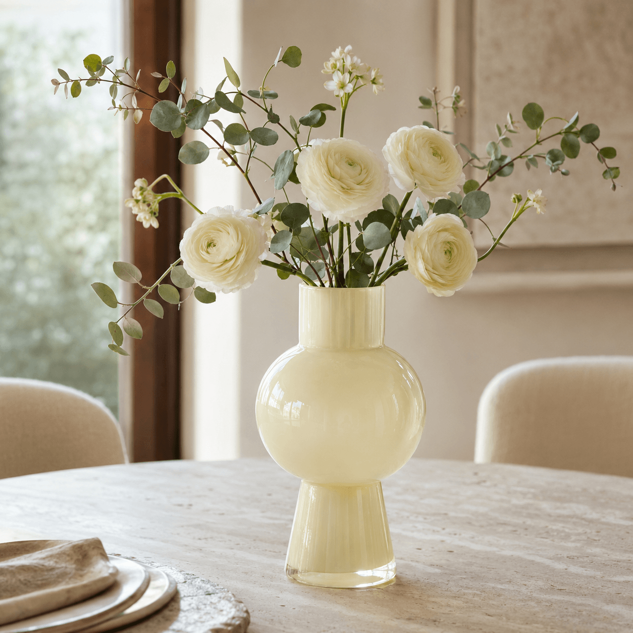 Tall ivory glass vase holding white ranunculus and eucalyptus on a limestone dining table