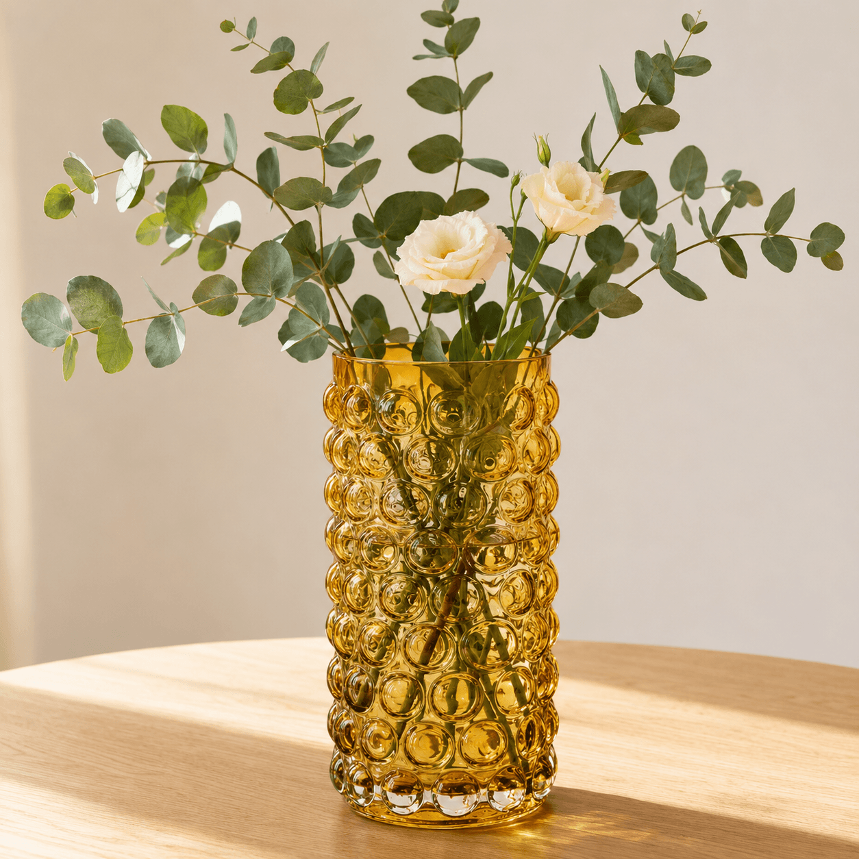 Amber tall bubble glass vase holding fresh eucalyptus branches and cream flowers on a light wood table