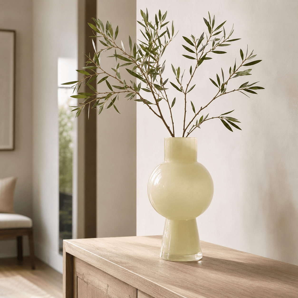 Tall ivory glass vase holding airy olive branches on a pale wood entryway console