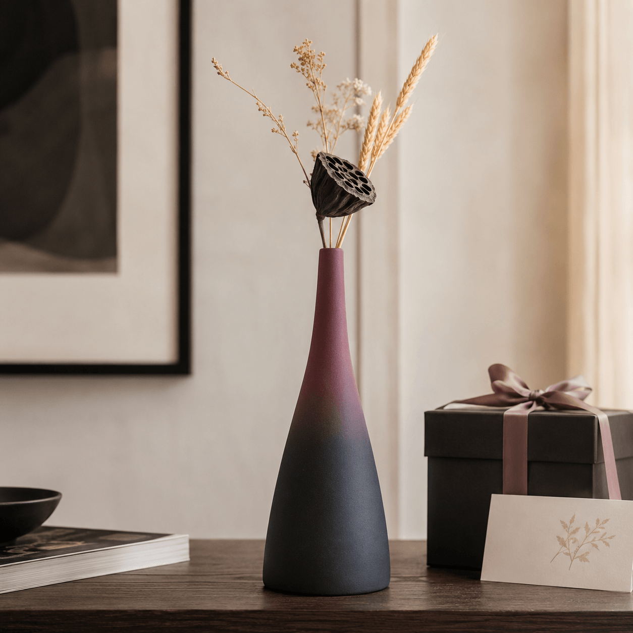 Slim tall mauve and charcoal ceramic vase with dried stems beside a gift box
